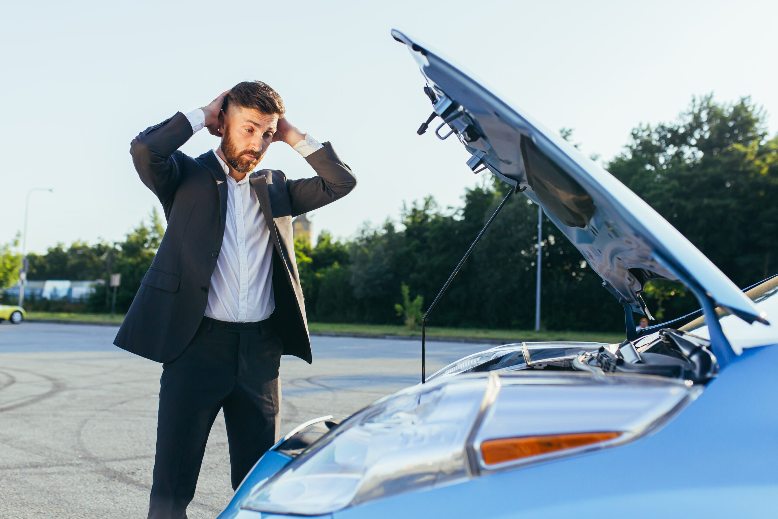 A frustrated man standing next to a broken-down car, using his phone to call for roadside assistance and insurance help.