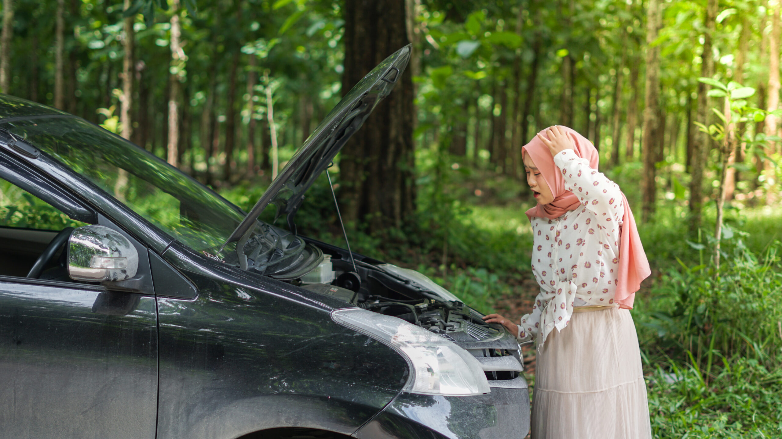 A young Asian woman wearing a hijab stands next to her car with the hood open, looking concerned about a possible breakdown.