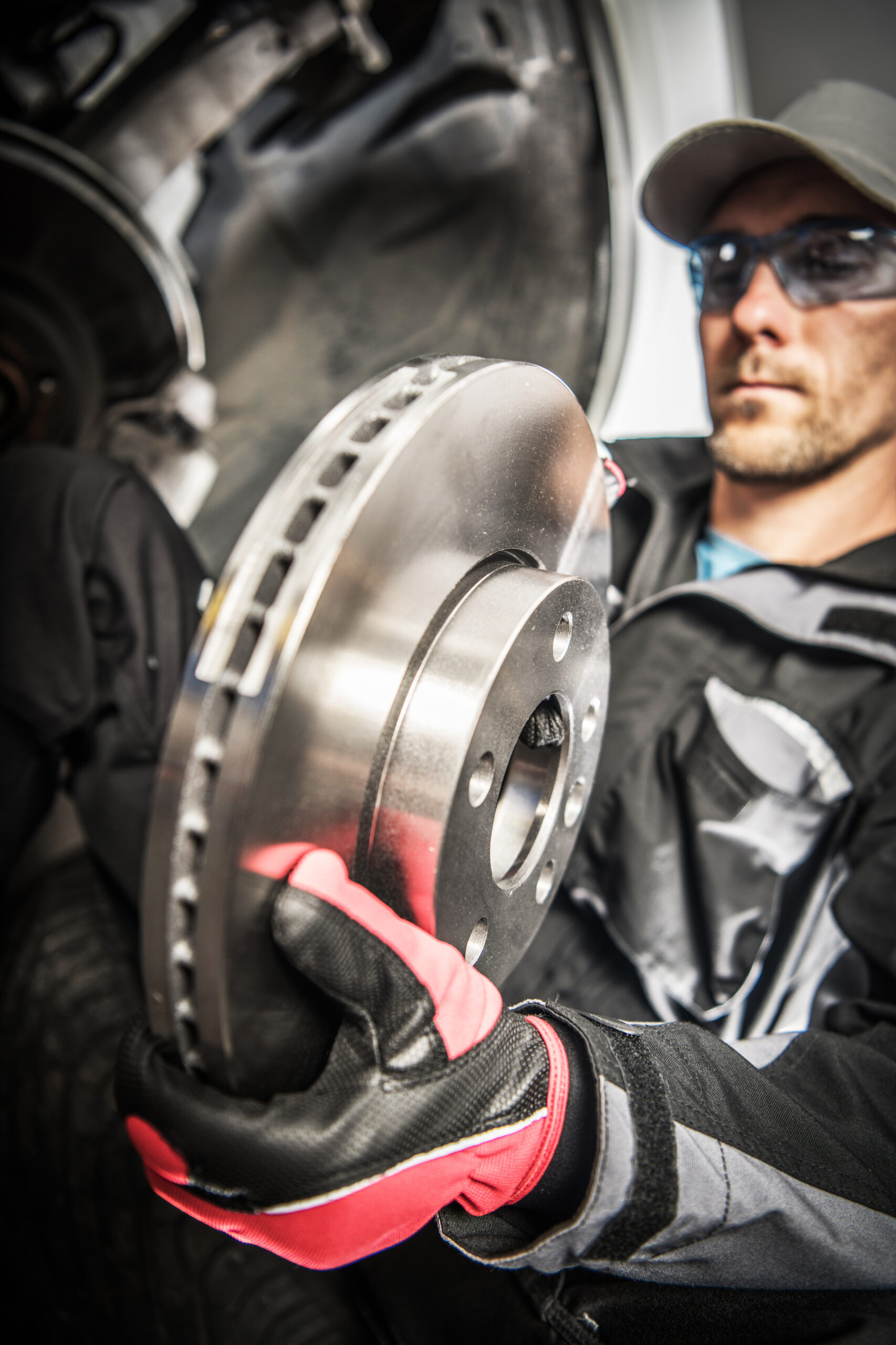 A mechanic in a repair shop, wearing safety glasses and gloves, carefully removes a worn brake disc from a car's wheel hub.