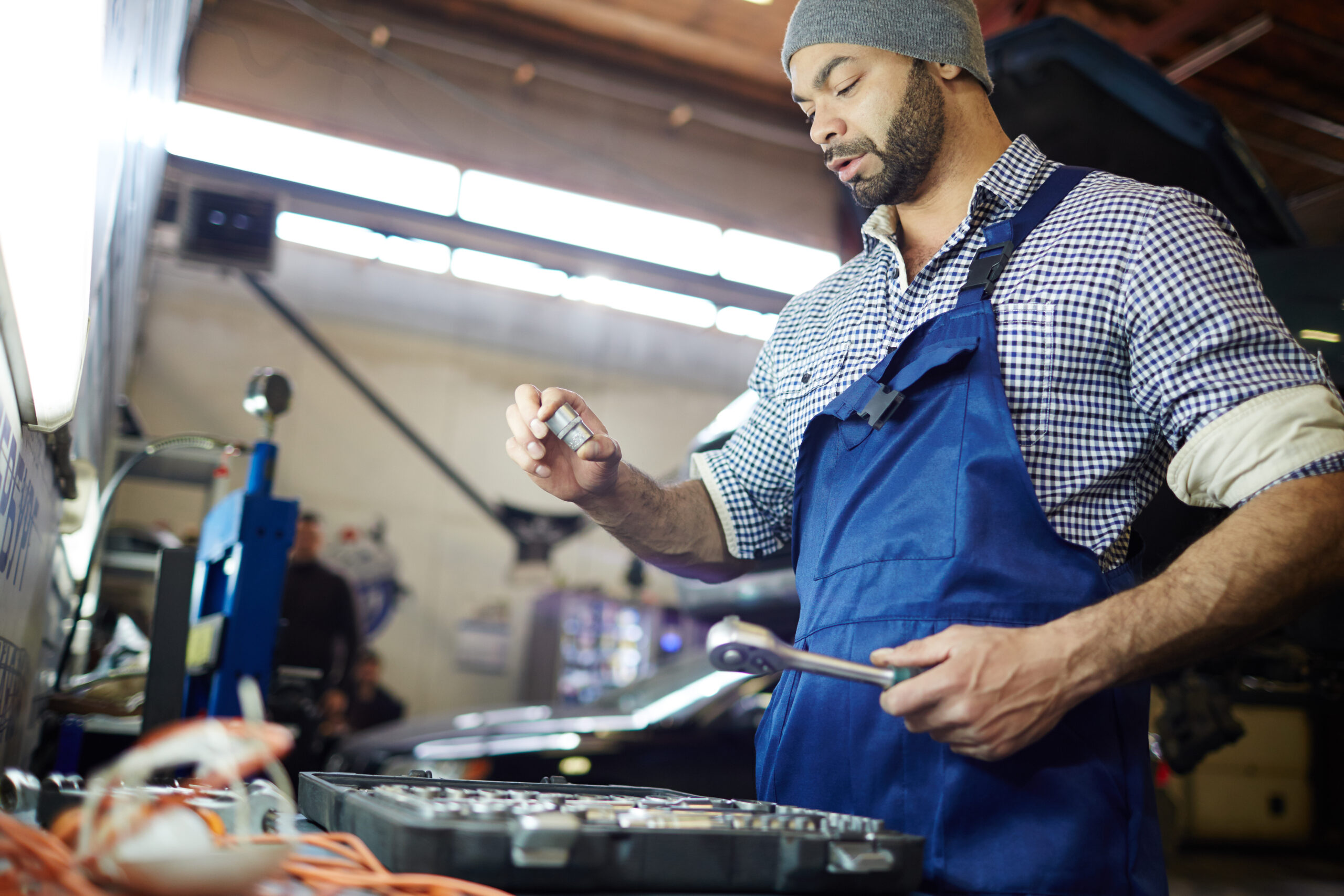 A car repairman in uniform leans over an open car hood, holding a wrench.