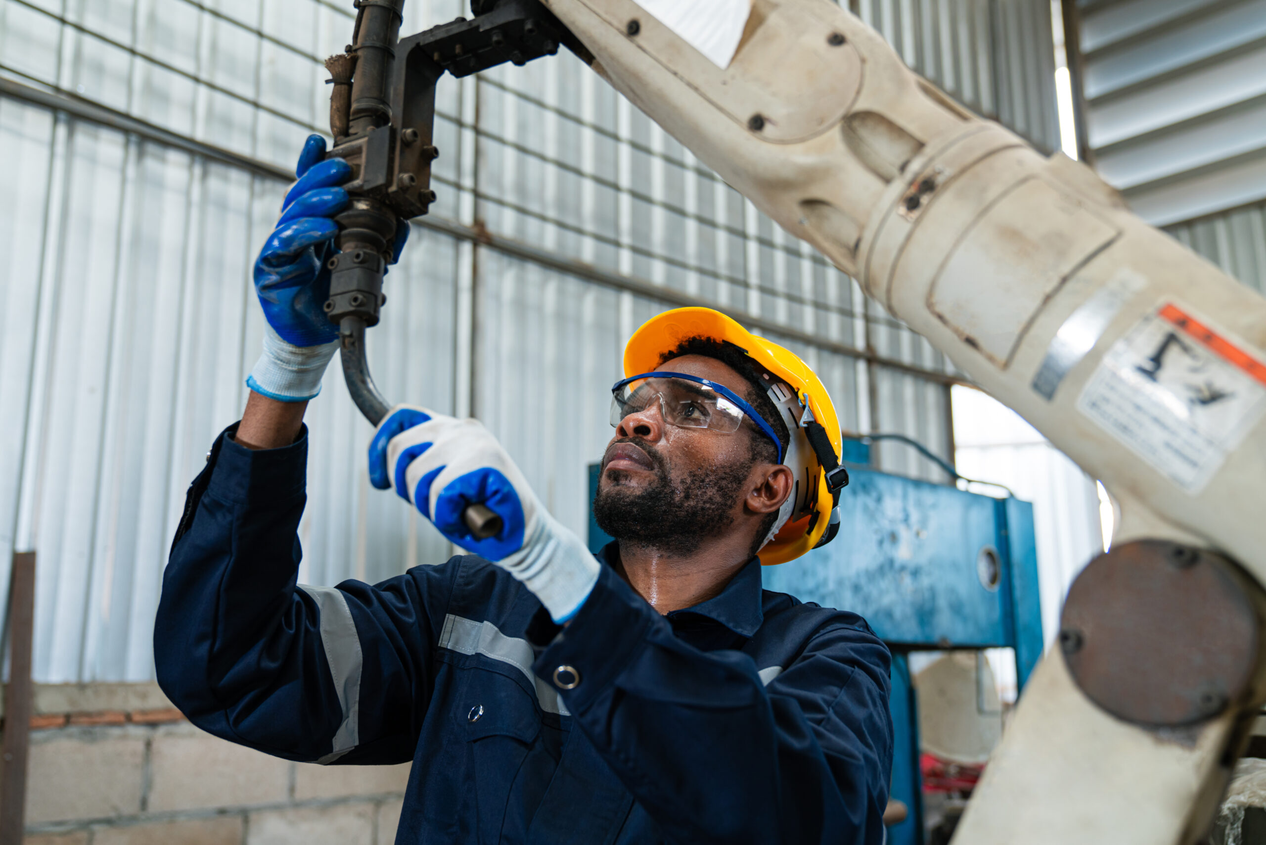 A male mechanic, wearing a blue uniform and safety glasses, carefully inspects and repairs a car's exhaust pipe in a well-equipped garage.