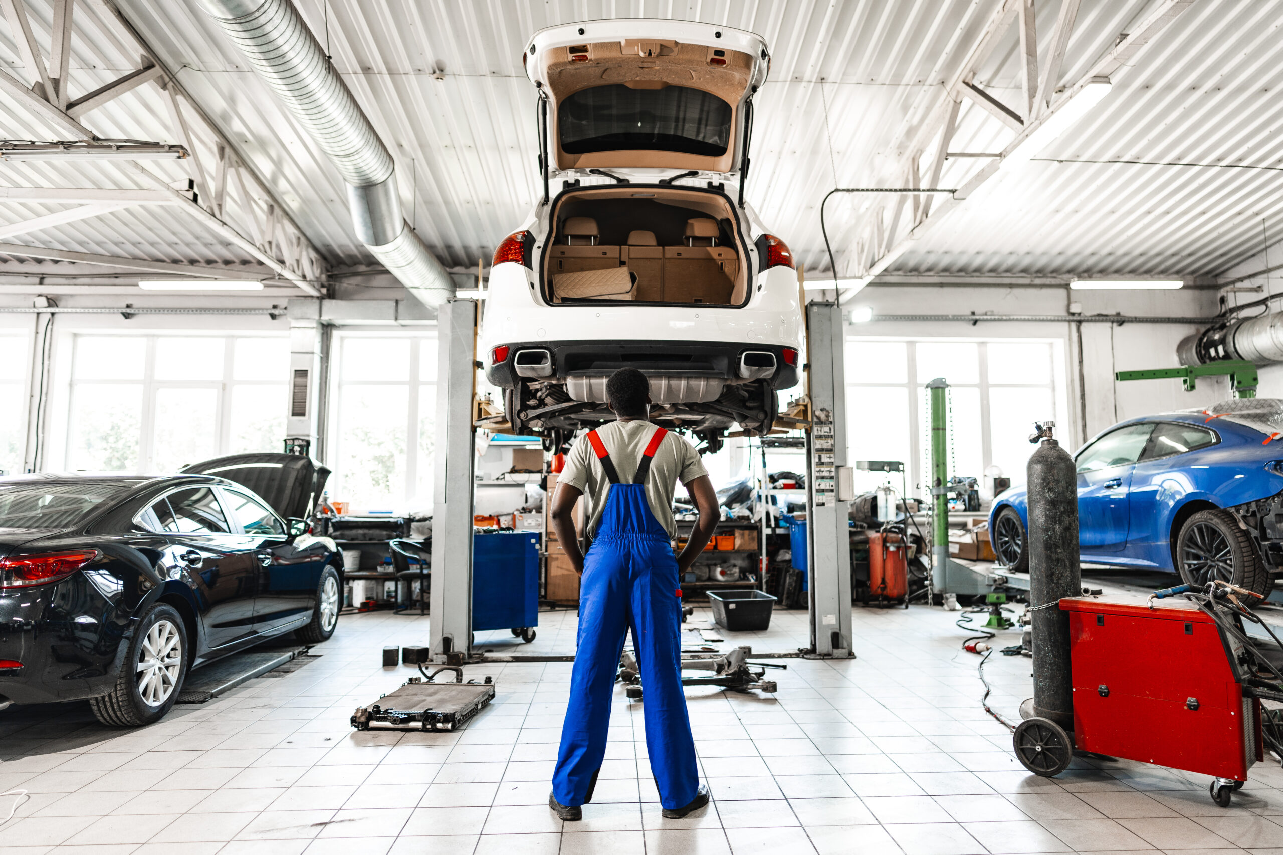 A young African mechanic with a thoughtful expression stands in a car service shop, looking at a vehicle's engine.