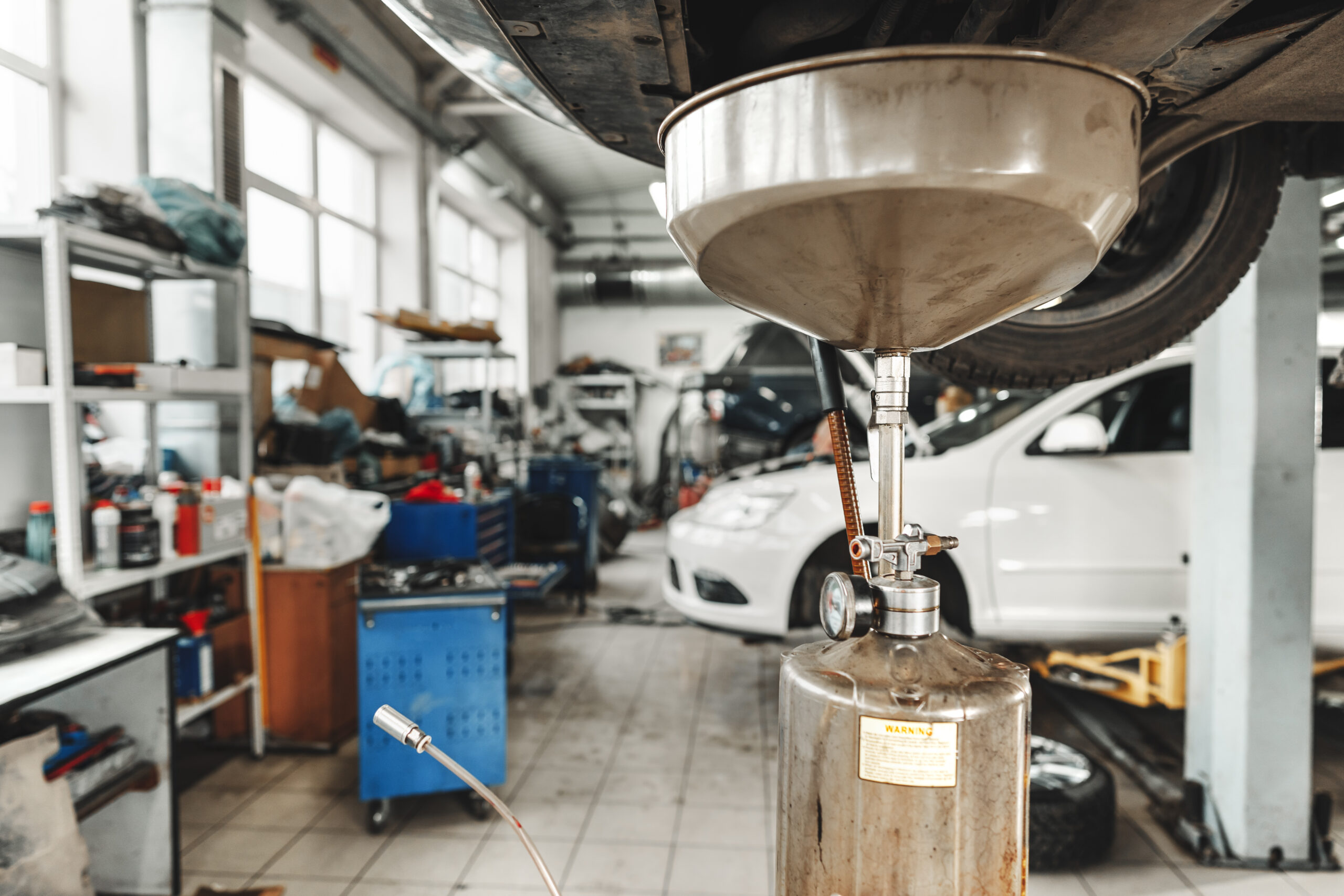 A mechanic in a garage, wearing gloves, drains old oil from a car's engine into a drain pan.