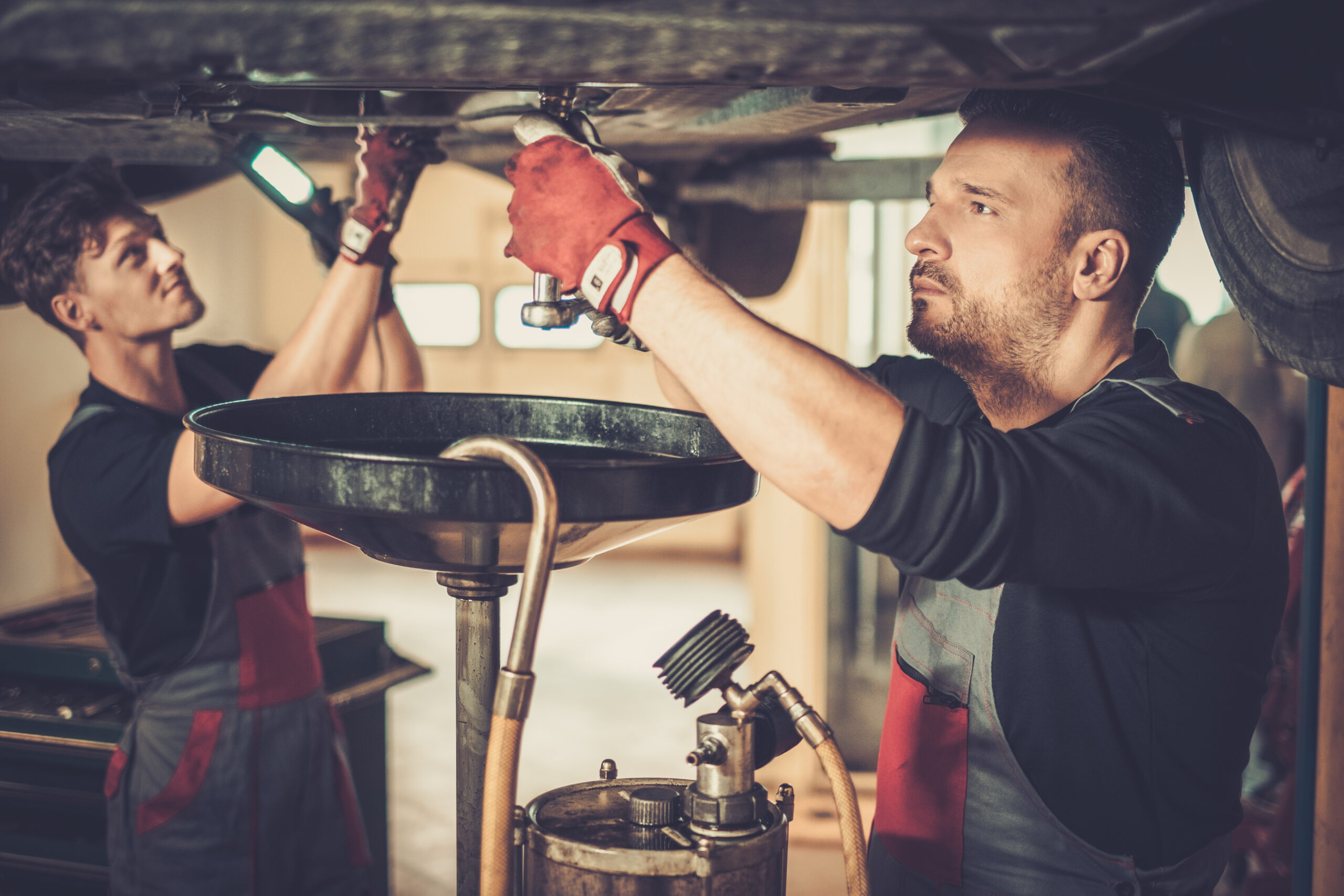 A professional car mechanic wearing a blue uniform carefully drains used motor oil from a car's engine into a pan.