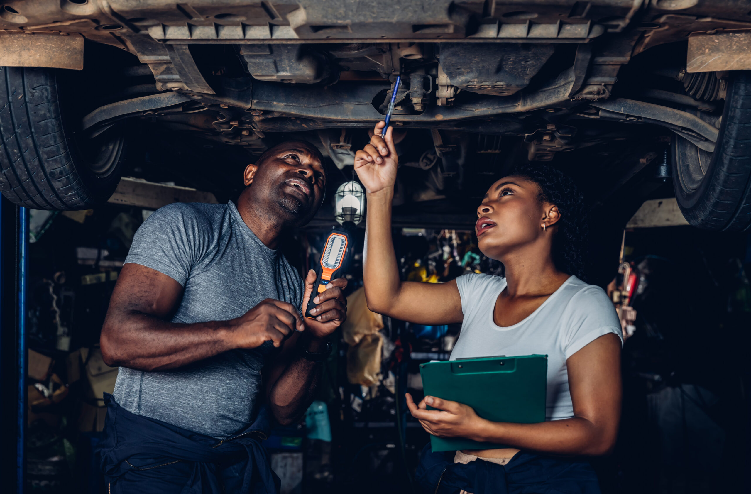 Experienced mechanic in work overalls using a flashlight to inspect the underside of a car raised on a lift.