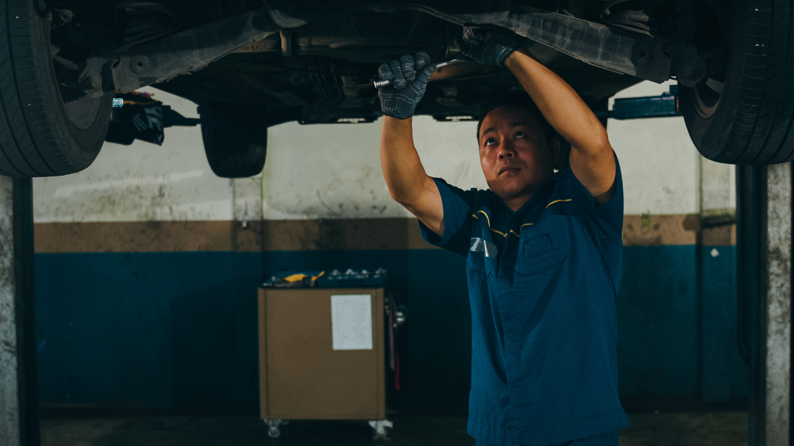 A mechanic in coveralls carefully tightening bolts on a car engine raised on a lift.