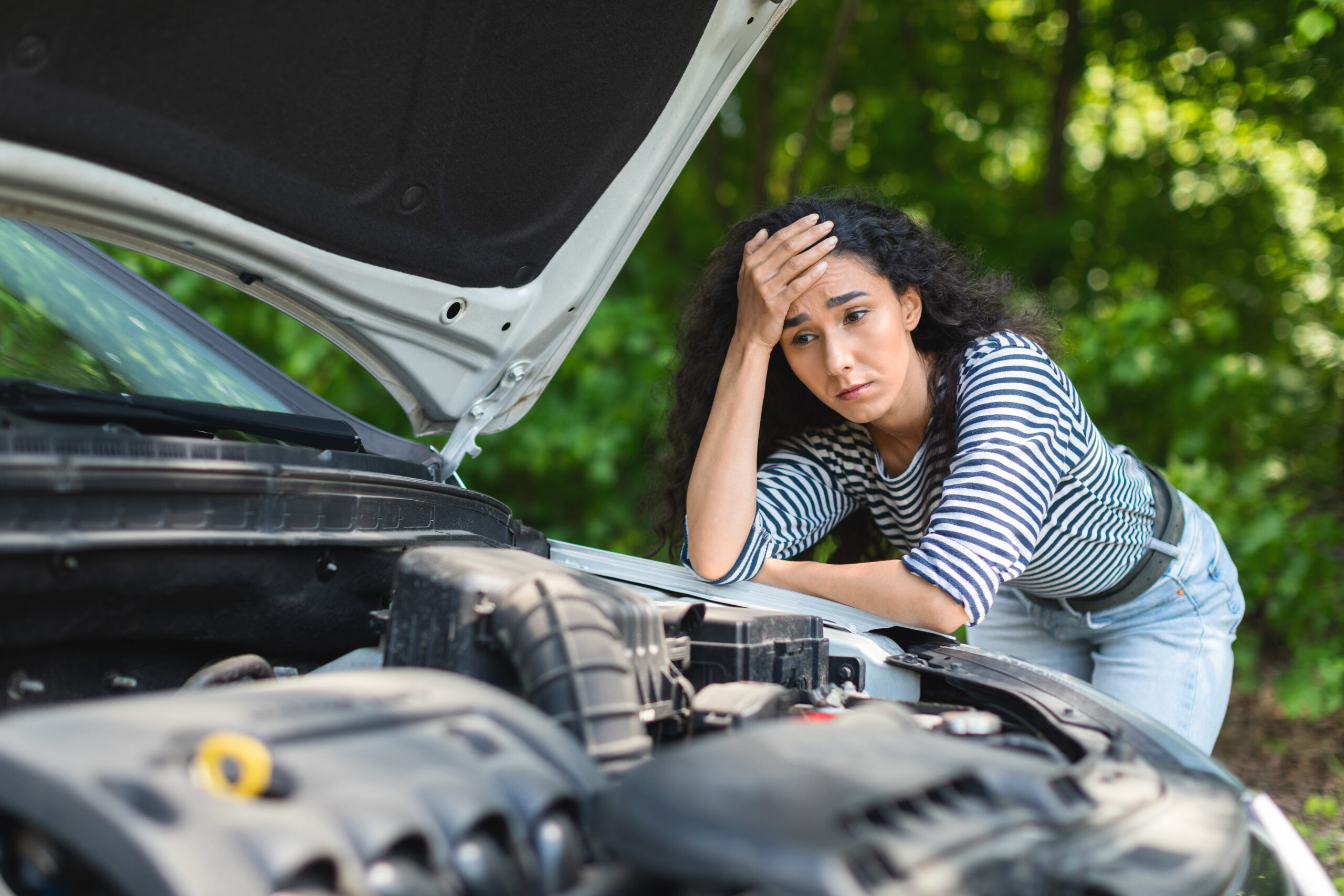 A disheartened brunette woman leans on her car's open hood, awaiting roadside assistance.