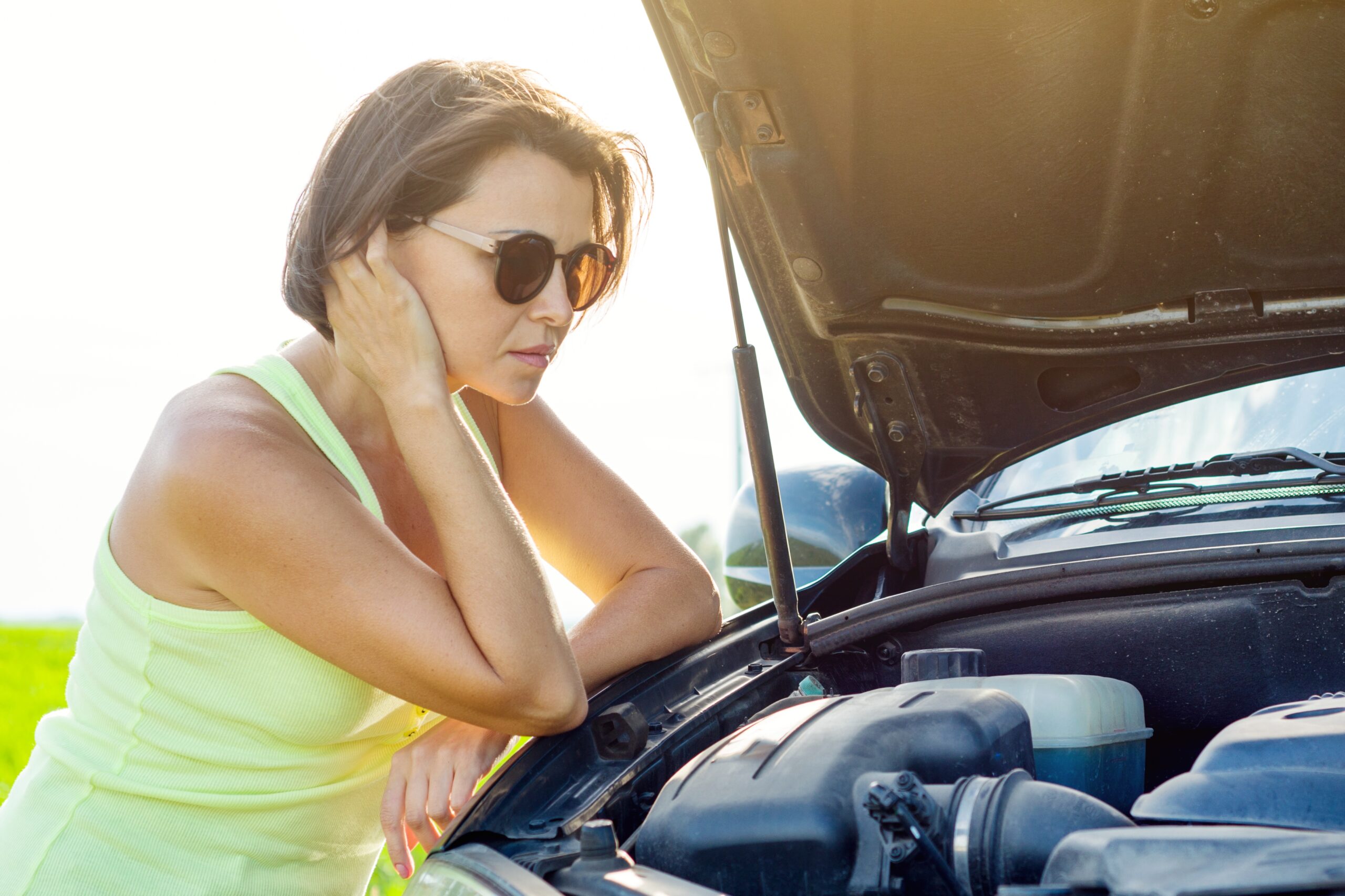 A young woman, looking distressed, stands beside her broken-down car on a rural road.