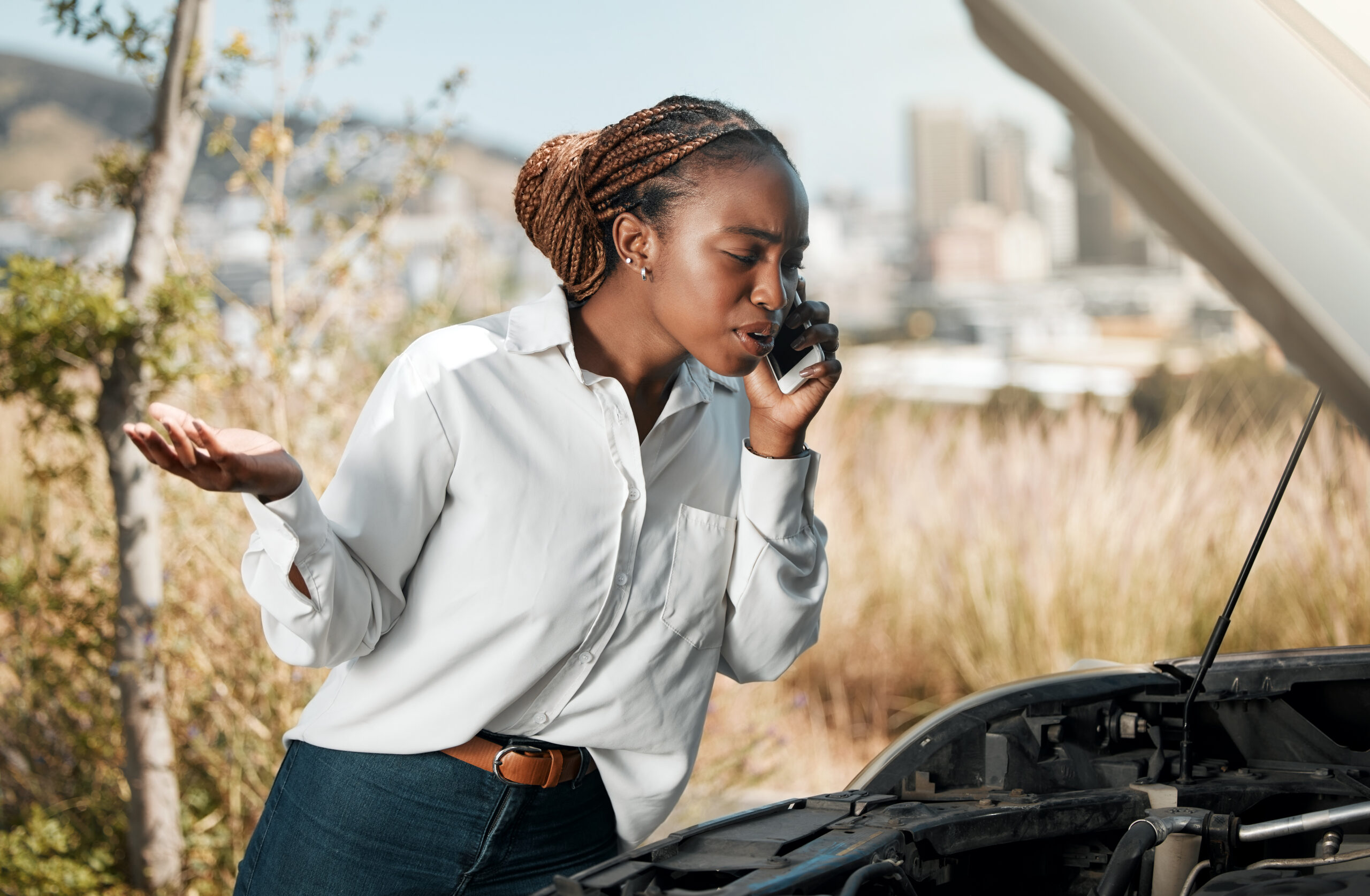 A Black woman with a frustrated expression on her face makes a phone call while standing next to her broken-down car on the side of the road.