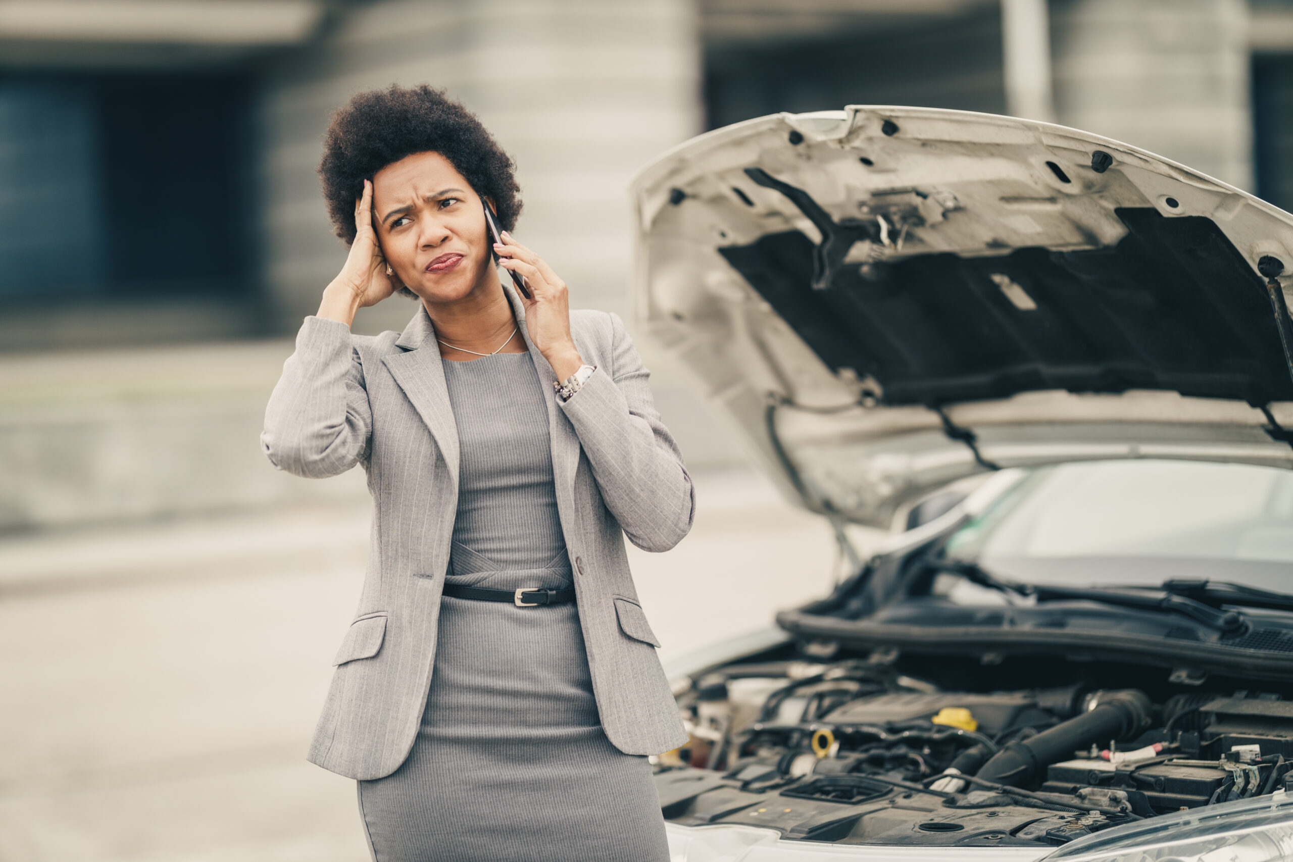 A frustrated driver with their head in their hands, standing next to a car with the hood up.