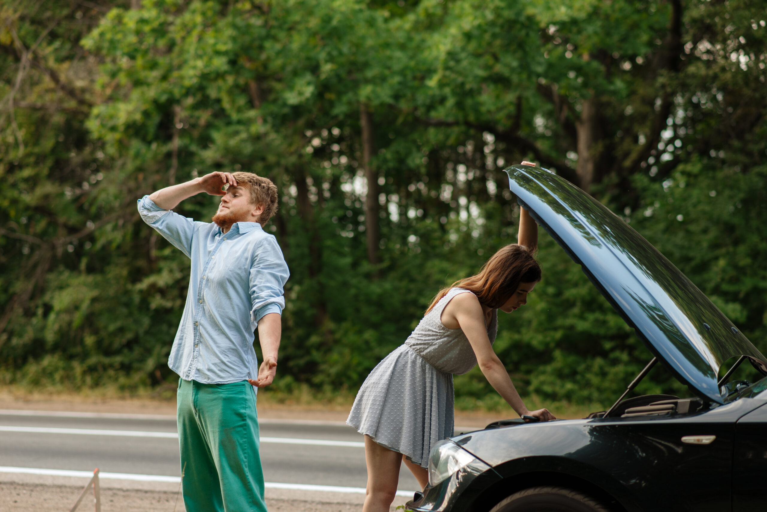 A frustrated couple standing next to their car with the hood open, stranded on the side of the road.