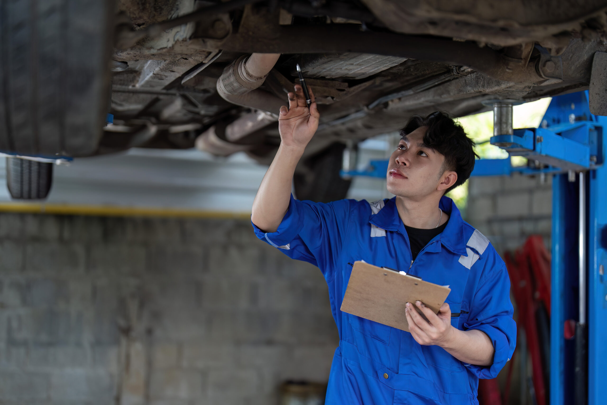 Asian mechanic inspecting the undercarriage of a car during routine maintenance.