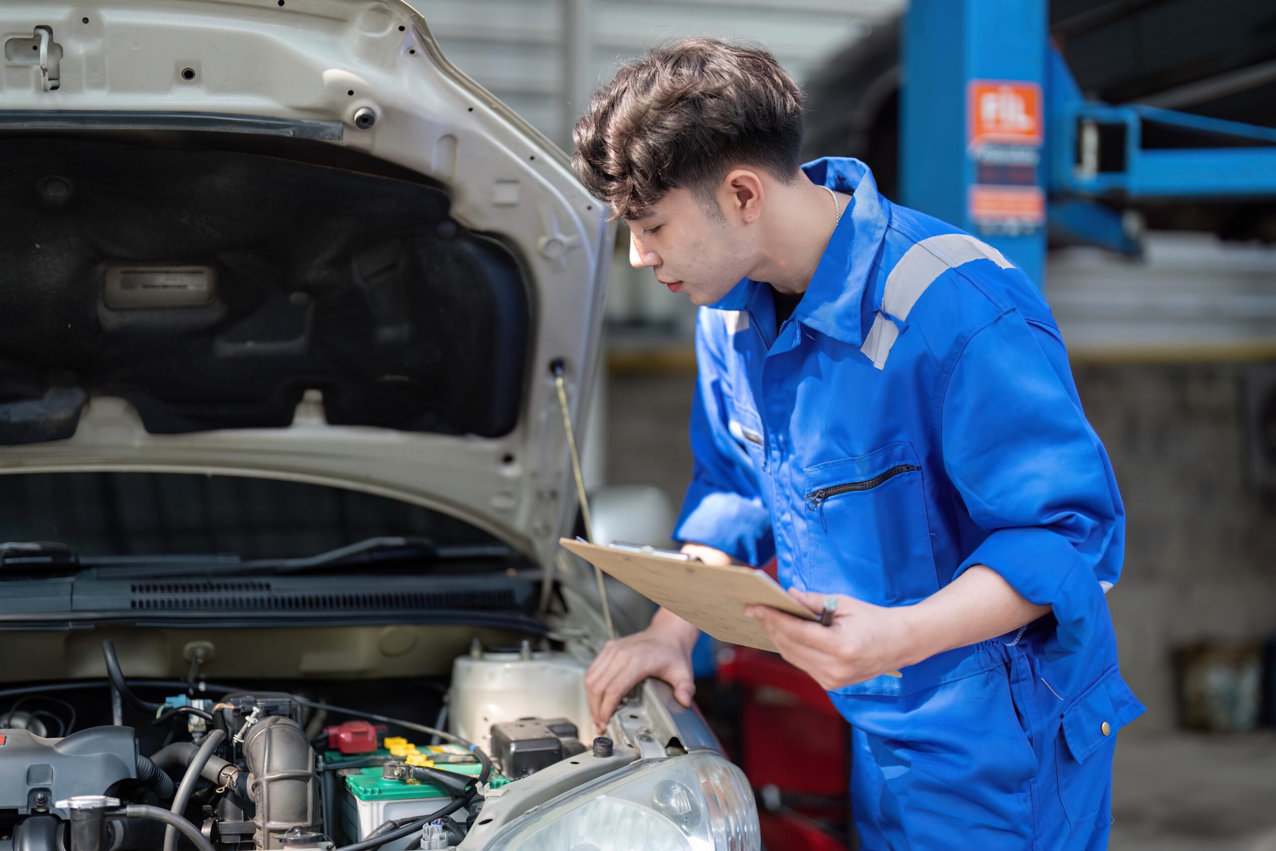 Asian mechanic inspecting car underside with checklist in garage.