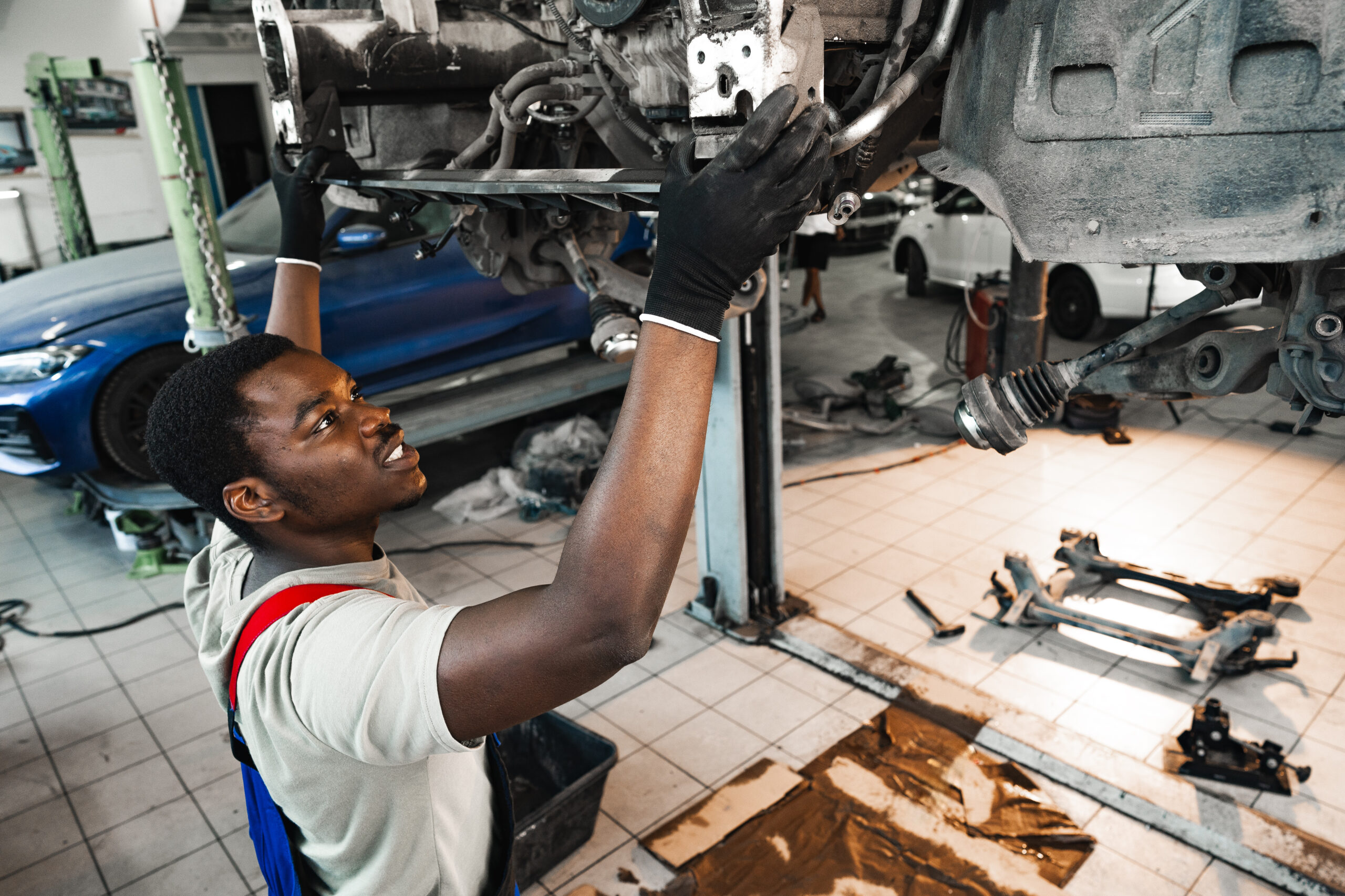 A young African mechanic in a blue uniform, lying on a creeper under a car in a brightly lit service center, carefully inspecting the undercarriage.